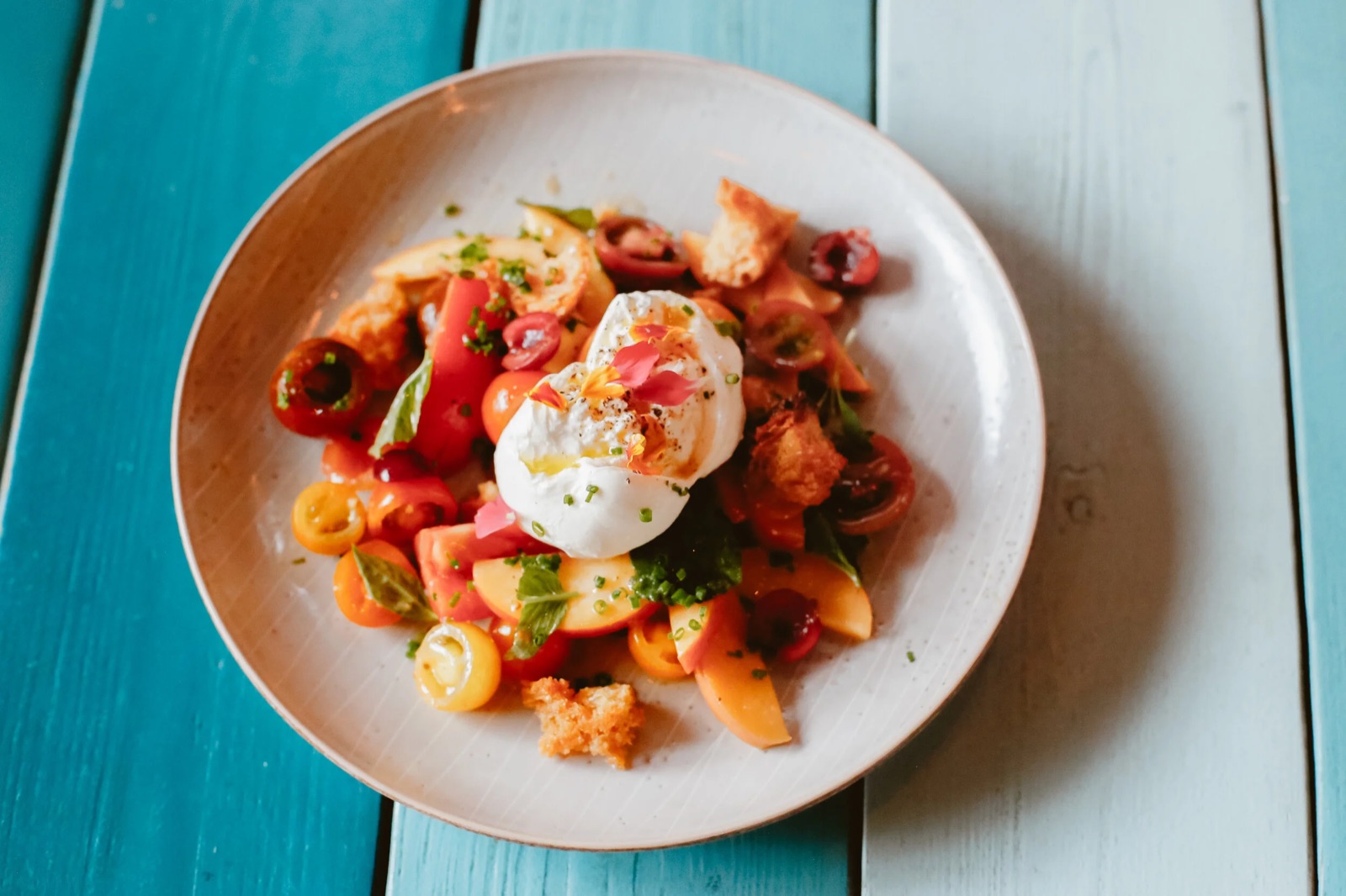 A plate of grilled peach and tomato salad with stracciatella on a teal table